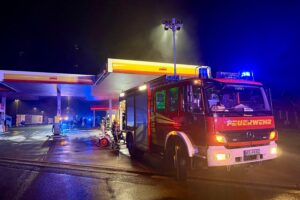 Brennender Pkw an Tankstelle, Feuerwehr im Einsatz, nächtliche Szene mit Rauch und Flammen.
