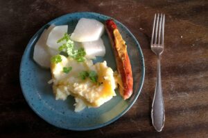 Bratwurst mit Kartoffelbrei und Kohlrabi auf einem Teller, symbolisiert regionale, preisbewusste Ernährung.