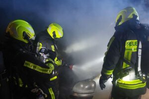 Zwei brennende PKW, Feuerwehr im Einsatz mit Löschschläuchen und Atemschutzgeräten.