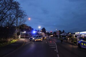 Ein Unfallort mit einem beschädigten Auto, Einsatzkräften und einer abgesperrten Straße.