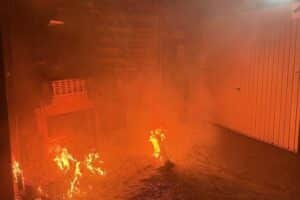 Ein brennender Anbau mit Feuerwehr im Einsatz und rauchendem Einfamilienhaus im Hintergrund.
