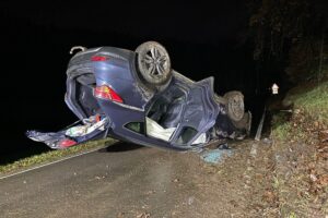 Ein überschlagenes Auto liegt auf dem Dach, Unfallstelle auf einer ländlichen Straße.