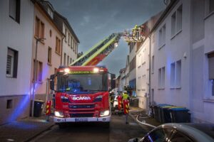 Brand im Mehrparteienhaus, unbewohnbar, Feuerwehr im Einsatz, Rauchsäule und zerstörte Dachstruktur.