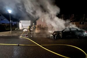 Ein brennender Pkw und Feuerwehrleute im Einsatz, um das Feuer zu löschen.