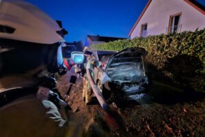 Feuerwehrleute bekämpfen einen brennenden Pkw, Flammen greifen auf Hecke über.