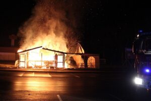 Flammen und Rauch: abgebrannter Imbiss auf Parkplatz, Feuerwehr löscht, Polizei ermittelt Brandursache.