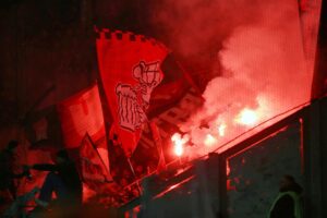 Fans im Stadion zeigen Pyrotechnik, während Sicherheitsmaßnahmen und Gewaltproblematik diskutiert werden.