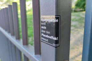 Aufkleber gegen Homöopathie auf einem Plakat, mit politischem Hintergrund und gesundheitlichen Argumenten.