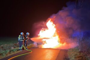 Ein ausgebrannter Transporter mit Feuerwehrleuten, die Löscharbeiten durchführen.
