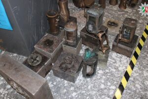 Drei Männer stehen vor Grabsteinen, umgeben von gestohlenen Bronze- und Metallgrablichtern.