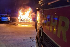 Zwei brennende Fahrzeuge auf einem Parkplatz, Feuerwehrleute löschen den Brand.