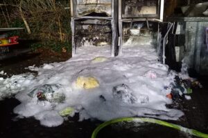 Brennende Altkleidercontainer nach Feuerwehrlöscharbeiten, Einsatzkräfte bei der Brandbekämpfung sichtbar.