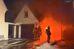 Feuerwehr im Einsatz: Auto brennt unter Carport, Nachbarn werden geschützt. Winterliche Bedingungen sichtbar.
