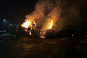 Feuerwehr beim Löschen eines Brandes in der Silvesternacht, Einsatzfahrzeuge und brennende Hecken sichtbar.