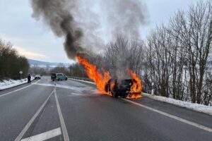 PKW in Flammen auf Landstraße, Feuerwehr im Einsatz, Rauch und Zerstörung sichtbar.