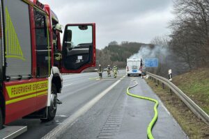 Brennendes Wohnmobil auf Autobahn, Feuerwehr im Einsatz, Flammen und Rauch sichtbar.