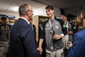 Friedrich Merz gratuliert Handballern persönlich nach EM-Finale, umarmt Spieler und posiert für Gruppenfoto.