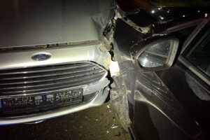Unfallaufnahme mit zwei beschädigten Fahrzeugen und Polizisten, Rettungswagen im Hintergrund, nächtliche Straße.