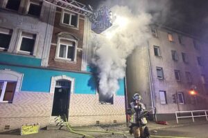 Brand in Mehrfamilienhaus: Feuerwehr rettet 14 Personen, Flammen schlagen aus Fenster, dramatische Szene.