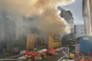 Feuerwehr löscht brennenden Müllcontainer vor Klinikum, starke Rauchentwicklung und geschützte Gebäude.