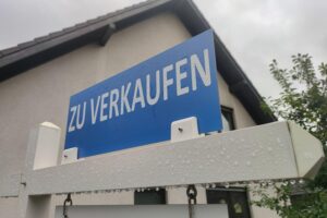 Ein "Zu Verkaufen"-Schild vor einem Immobilienobjekt, symbolisiert die Steuerproblematik in Deutschland.
