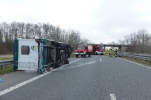 Ein überschlagenes Wohnmobil auf der Autobahn, mit Polizei und Rettungswagen im Hintergrund.