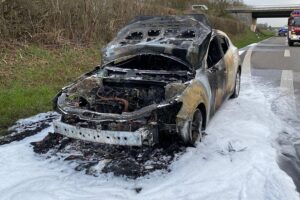 Ein ausgebrannter PKW auf der Autobahn mit Feuerwehr und Verkehrsbehinderungen im Hintergrund.