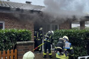 Feuerwehr löscht Dachstuhlbrand; Rauchsäulen, Feuerwehrfahrzeuge und Einsatzkräfte vor einem Wohnhaus.
