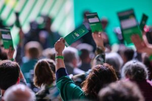 Abstimmung auf einem Grünen-Parteitag mit engagierten Mitgliedern und farbenfrohen Banner im Hintergrund.