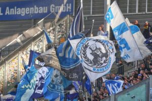 Fans beider Teams im Stadion, friedliche Stimmung, Feuerwerkskörper und Polizeipräsenz im Hintergrund.