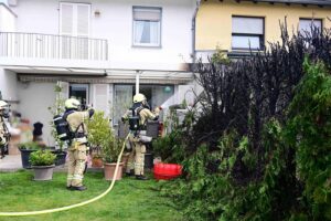 Brennende Hecke zwischen Reihenhäusern, Feuerwehr im Einsatz, Rauch und beschädigtes Wohnhaus sichtbar.