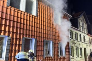 Feuerwehrlöschaktion bei Zimmerbrand in Mehrfamilienhaus, Rauch und evakuierte Personen sichtbar.