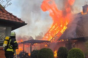 Feuerwehrleute bekämpfen intensiven Gebäudebrand; rauchende Trümmer und Löschfahrzeuge sind sichtbar.
