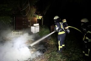 Feuerwehr löscht Flächenbrand im Wald, rettet Gartenhütten vor weiterer Ausbreitung.