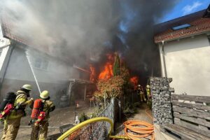 Feuerwehrleute bekämpfen Garagenbrand, Flammen bedrohen angrenzendes Wohnhaus, starke Rauchentwicklung sichtbar.