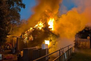 Brennende Gartenhütte mit Feuerwehr im Einsatz, Explosionsgefahr durch Gasflasche, dramatische Szene.