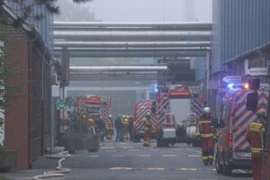 Feuerwehr im Einsatz bei Brand in Industriebetrieb, Flammen auf Dach und Rohrleitungen sichtbar.