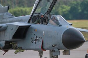 Zwei Politiker diskutieren über Rüstungsprojekte, während ein Kampfjet im Hintergrund sichtbar ist.