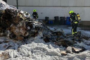 Müllpresse steht in Flammen, Feuerwehr im Einsatz, Rauch und Löschfahrzeug sichtbar.