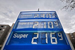 Tankstelle mit Preistafel, umrahmt von Menschen, die über hohe Energiepreise diskutieren.