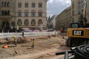 Bauarbeiten an einer Straße mit Baustellenfahrzeugen und Anwohnern, die besorgt zuschauen.