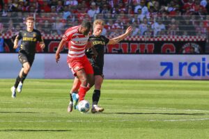 Zwei Fußballmannschaften stehen gegenüber, Fans jubeln, Spannung vor entscheidendem Spielmoment.