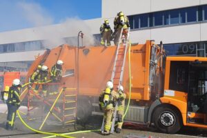 Rauchender Müllwagen im Wohngebiet, Feuerwehr im Einsatz zur Brandbekämpfung und Müllentleerung.