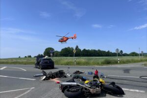 Rettungshubschrauber Christoph 13 über Unfallstelle, Einsatzkräfte bearbeiten verletzte Motorradfahrer und Pkw.