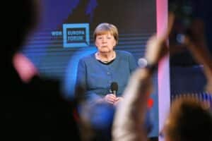 Angela Merkel spricht mit Ernst, umgeben von politischen Symbolen und einer dramatischen Kulisse.