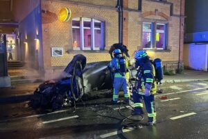 Feuerwehr löscht brennenden Pkw, verhindert Ausbreitung auf angrenzendes Gebäude. Zwei Personen betroffen.