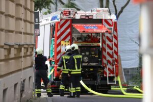 Einsatzkräfte der Feuerwehr löschen Brand in Neubaugebiet; Hecke, Wohnwagen und Pkw in Flammen.