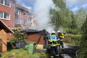 Holzschuppen brennt im Vorgarten, Feuerwehr im Einsatz, Flammen und Rauch sichtbar.