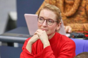 Ines Schwerdtner strahlt gelassen bei Parteitag, umringt von unterstützenden Genossen und diskutierenden Delegierten.