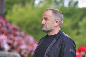 Manuel Baum lächelt selbstbewusst, umgeben von seinem Trainerteam beim FC Augsburg.